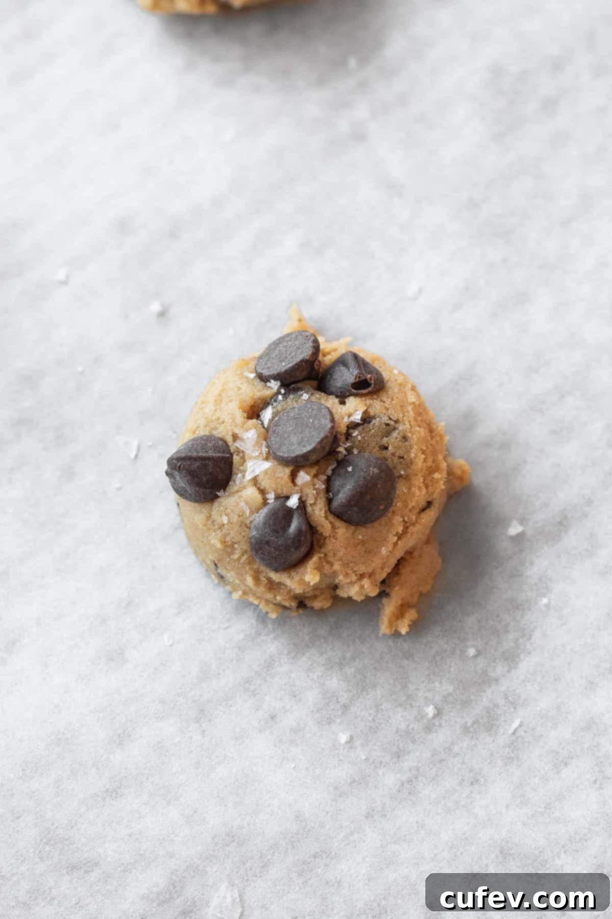 Close-up of a perfectly formed ball of gluten-free chocolate chip cookie dough, topped with extra chocolate chips and a sprinkle of coarse flaky salt, ready for baking.