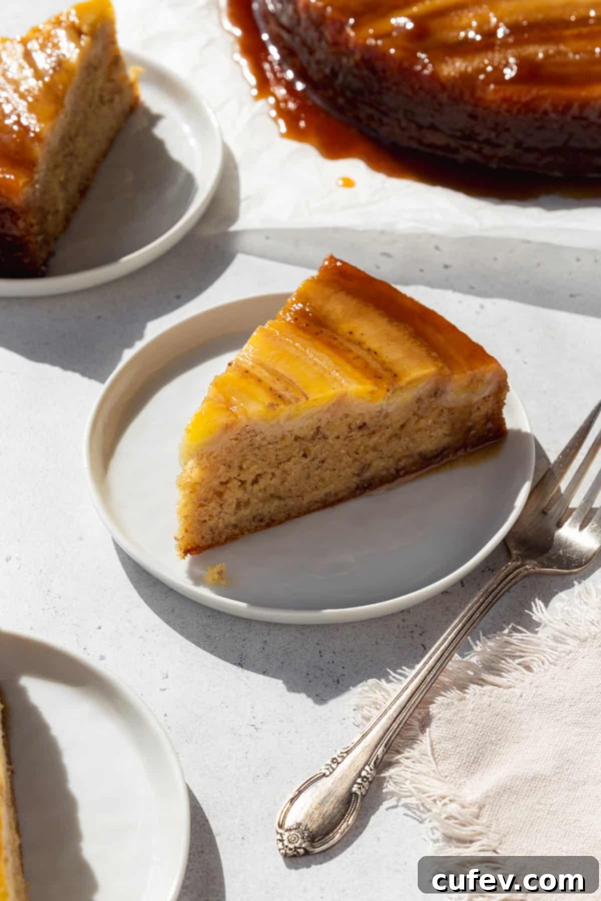 A slice of banana upside down cake on a white dessert dish with a silver fork next to it, showcasing the soft cake and sticky banana topping.