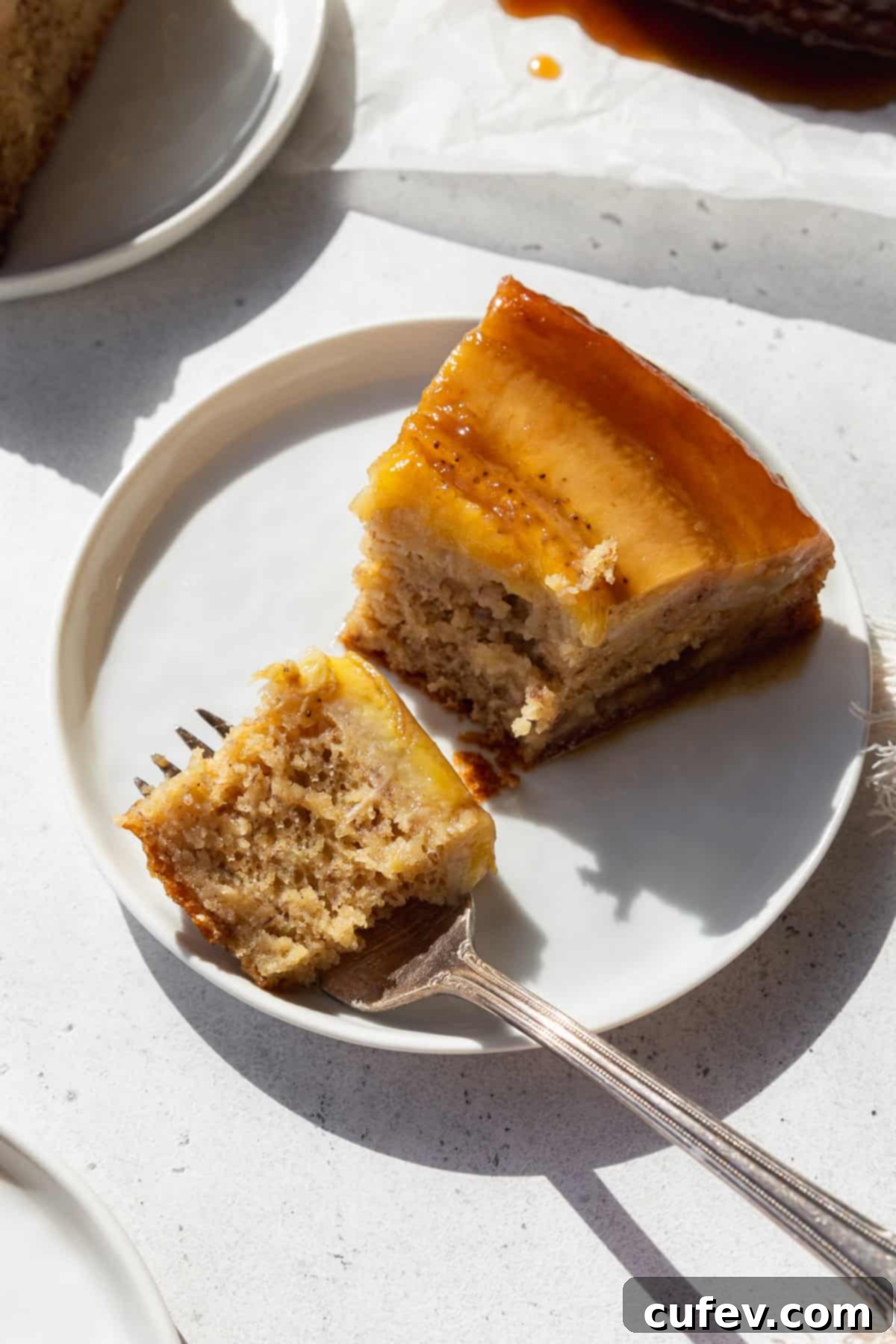 A slice of banana upside down cake on a white plate with a fork holding a bite of cake in front, highlighting the fluffy cake and gooey topping.