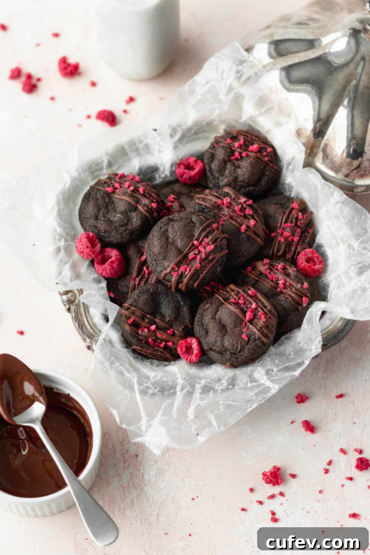 An overhead shot of perfectly round double chocolate chip cookies nestled in a silver dish lined with parchment paper, with freeze-dried raspberries scattered around for color and garnish.