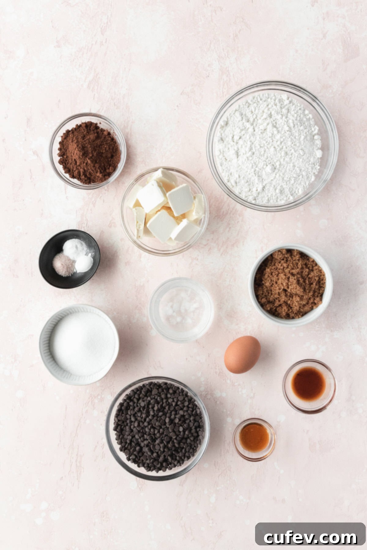 Ingredients for homemade double chocolate chip cookies, including butter, sugars, egg, honey, vanilla, flour, cocoa powder, salt, baking soda, baking powder, and chocolate chips, meticulously measured out into small bowls and arranged on a pink surface.