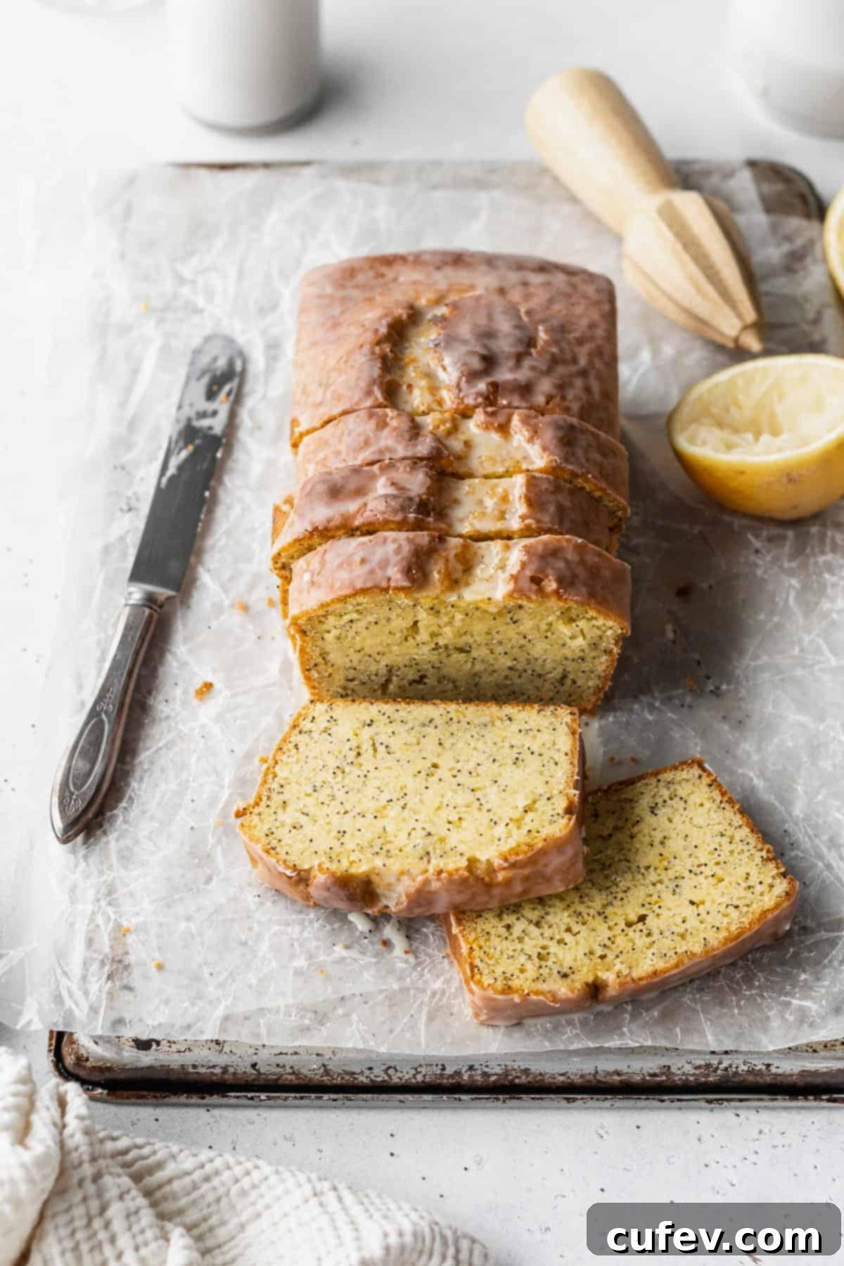 A sliced loaf of gluten-free lemon poppy seed loaf on a parchment paper lined baking sheet with a knife, half a lemon, and a wooden citrus zester placed around it.