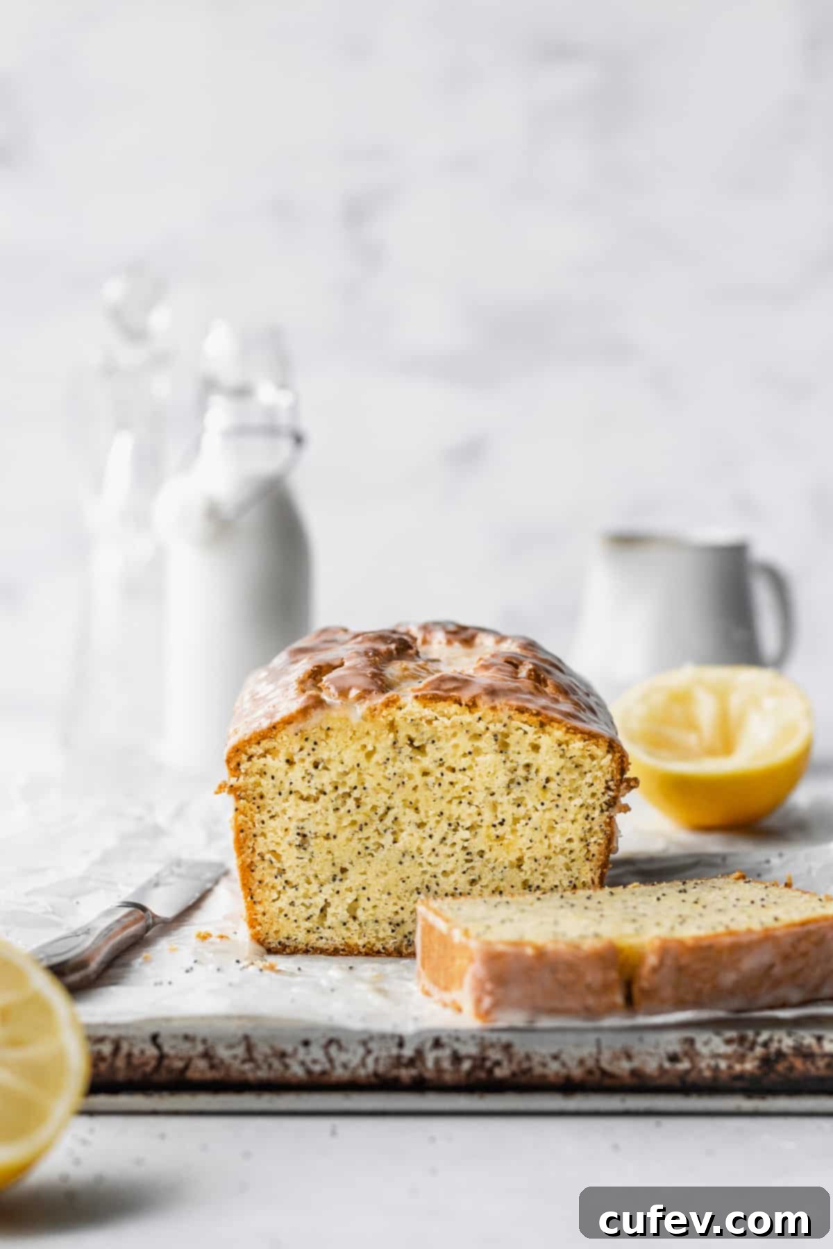 A sliced loaf of gluten free lemon poppy seed cake on a parchment paper with sliced lemons and milk jugs in the background.