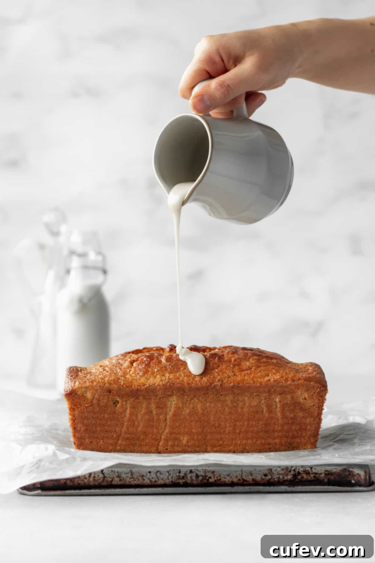A hand holding a ceramic jug of lemon icing being poured onto a loaf of gluten free lemon poppy seed cake.