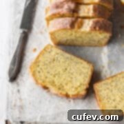 A slice of gluten free lemon poppy seed cake laying on parchment paper with a knife next to it and the sliced loaf behind it.