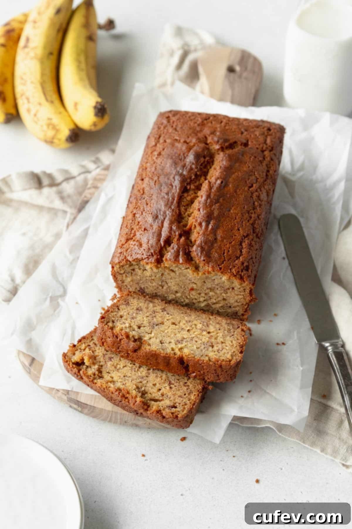 A sliced loaf of gluten free banana bread on sheets of parchment paper with a bunch of banana in the background.