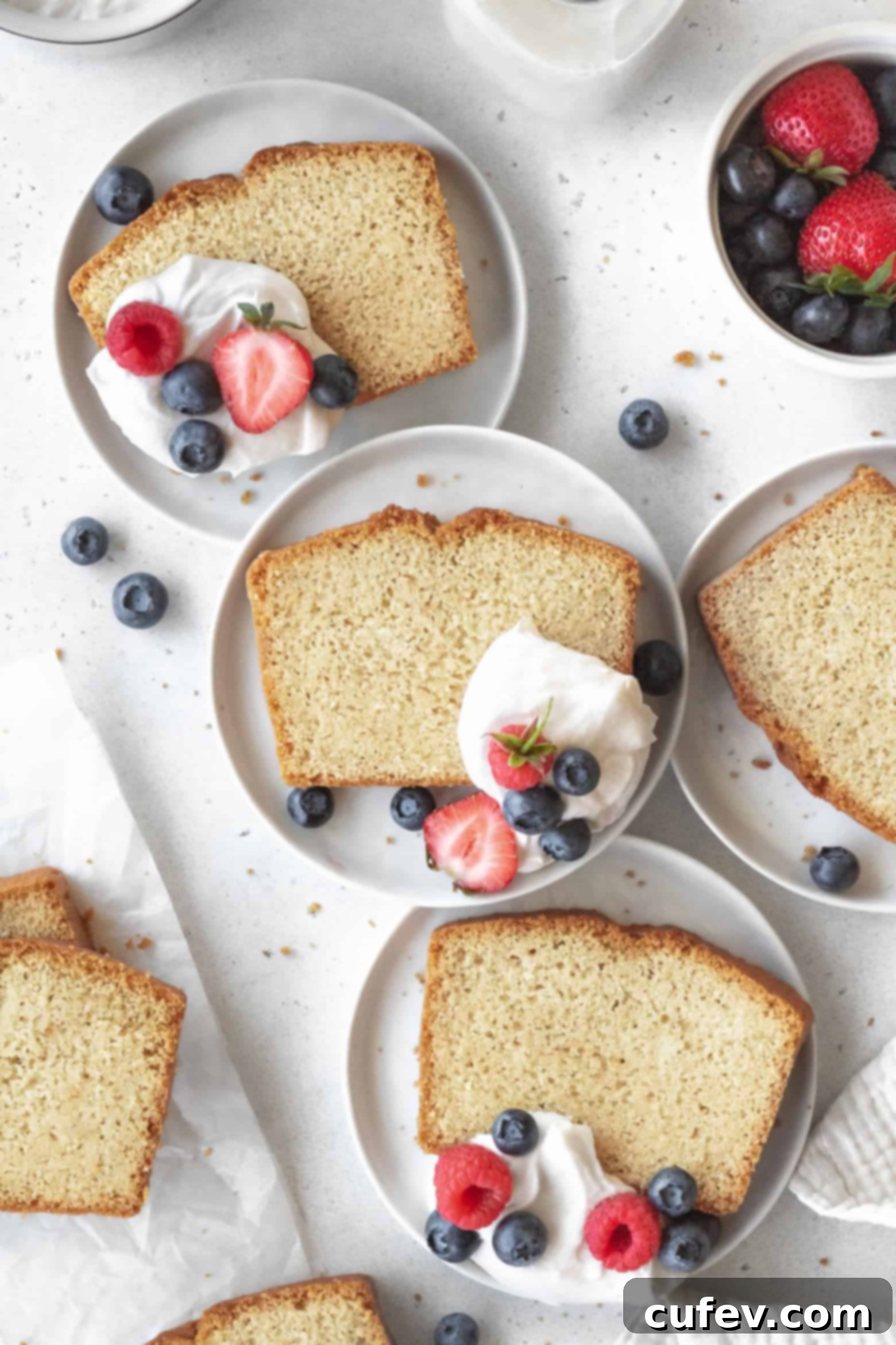 Multiple slices of tender gluten-free pound cake arranged on small white plates, accompanied by creamy coconut whipped cream and fresh berries.