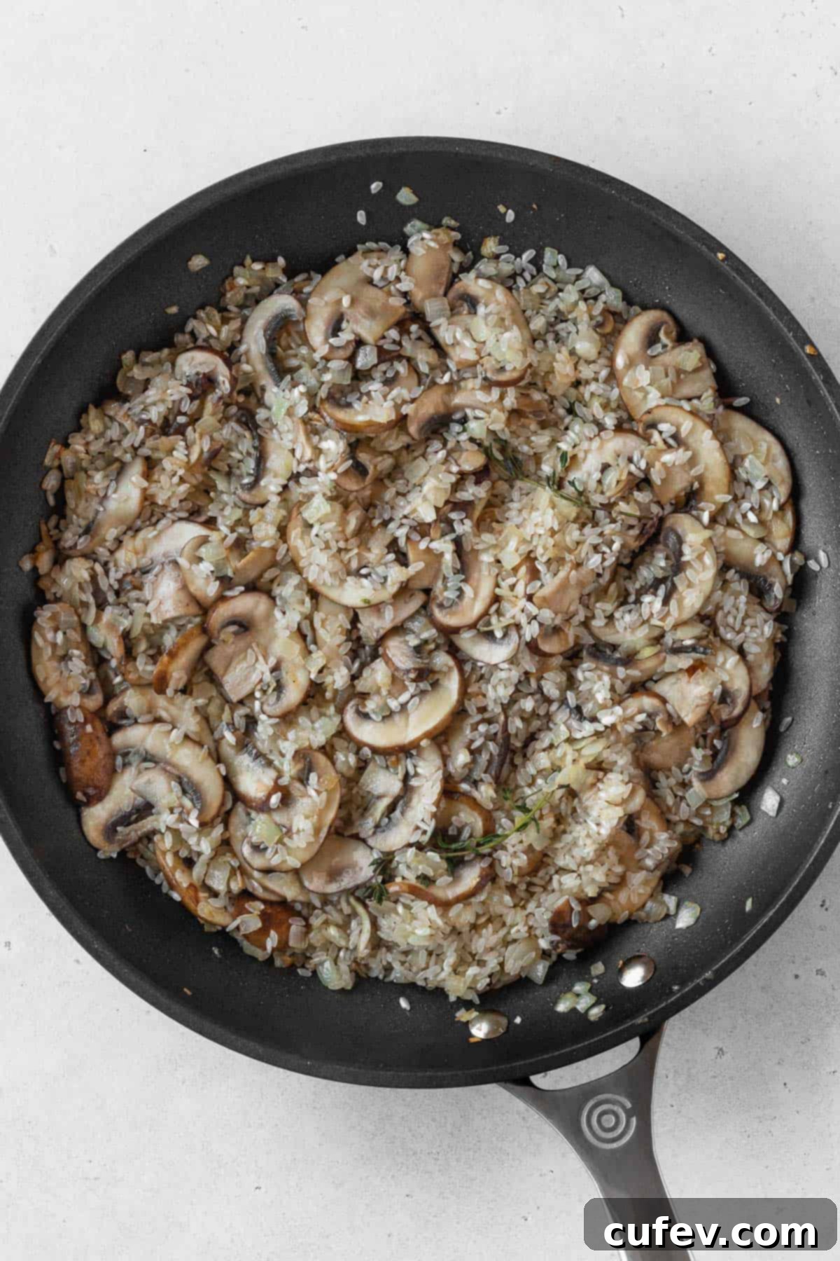 A close-up of onions, sliced mushrooms, and raw risotto rice gently toasting in a black non-stick pan.